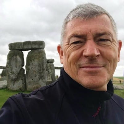 Person taking a selfie in front of the ancient Stonehenge monument under a cloudy sky.